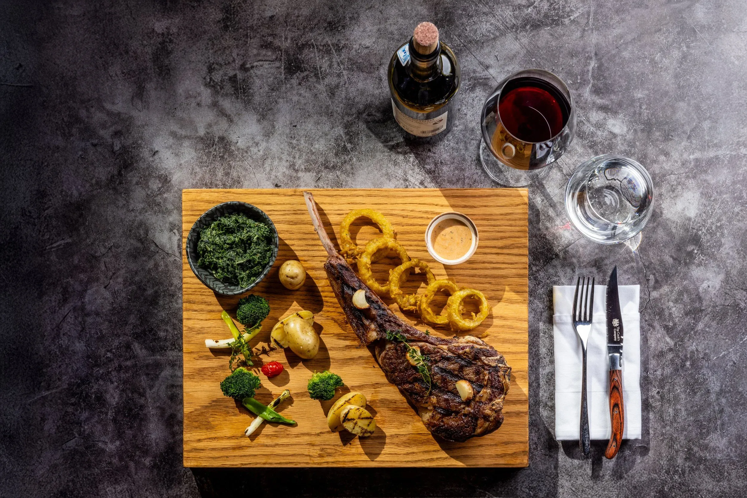 Overhead view of a tomahawk steak on a board with onion rings, sides and red wine